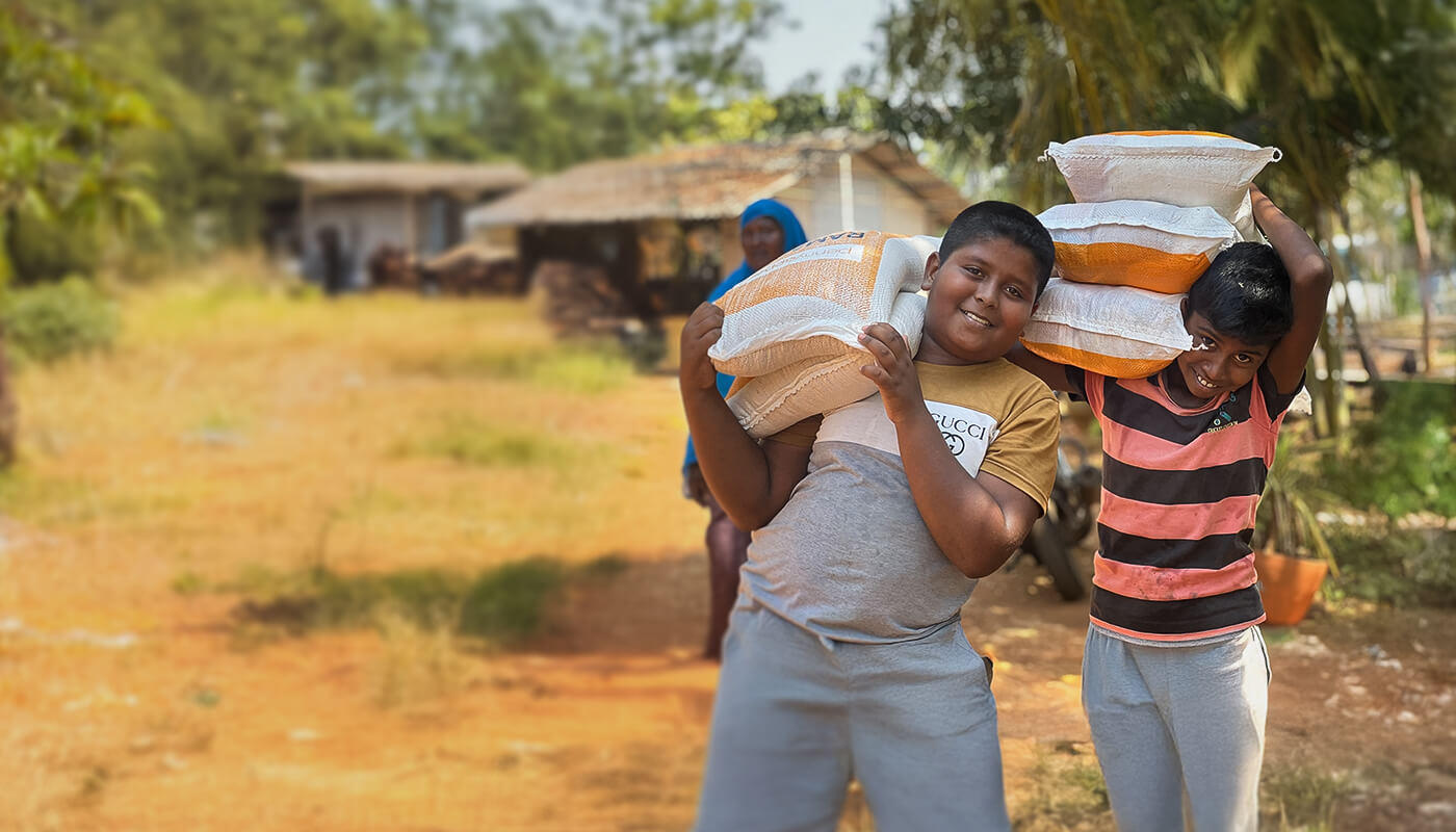 Two boys smile at the camera while carrying bags of food on their shoulders.