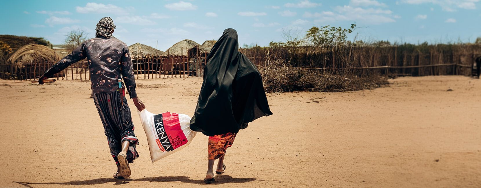 Two people walk across a dusty landscape, with one carrying a bag marked "KENYA." The background shows traditional huts and dry vegetation under a bright sky. One person is wearing a black niqab, while the other wears colorful attire.