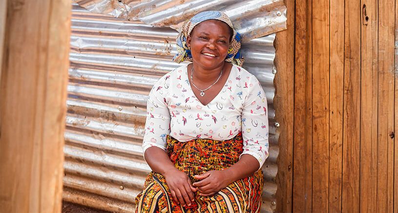 A joyful woman sitting on the ground outside a corrugated iron building. She is wearing a white patterned shirt and a colorful skirt, and she smiles warmly at the camera.