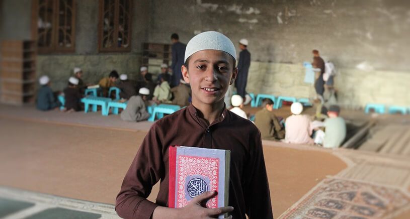 A young boy wearing a white prayer cap and traditional brown clothing smiles while holding a copy of the Quran. He stands inside a madrasa or Islamic school, with other children studying in the background.