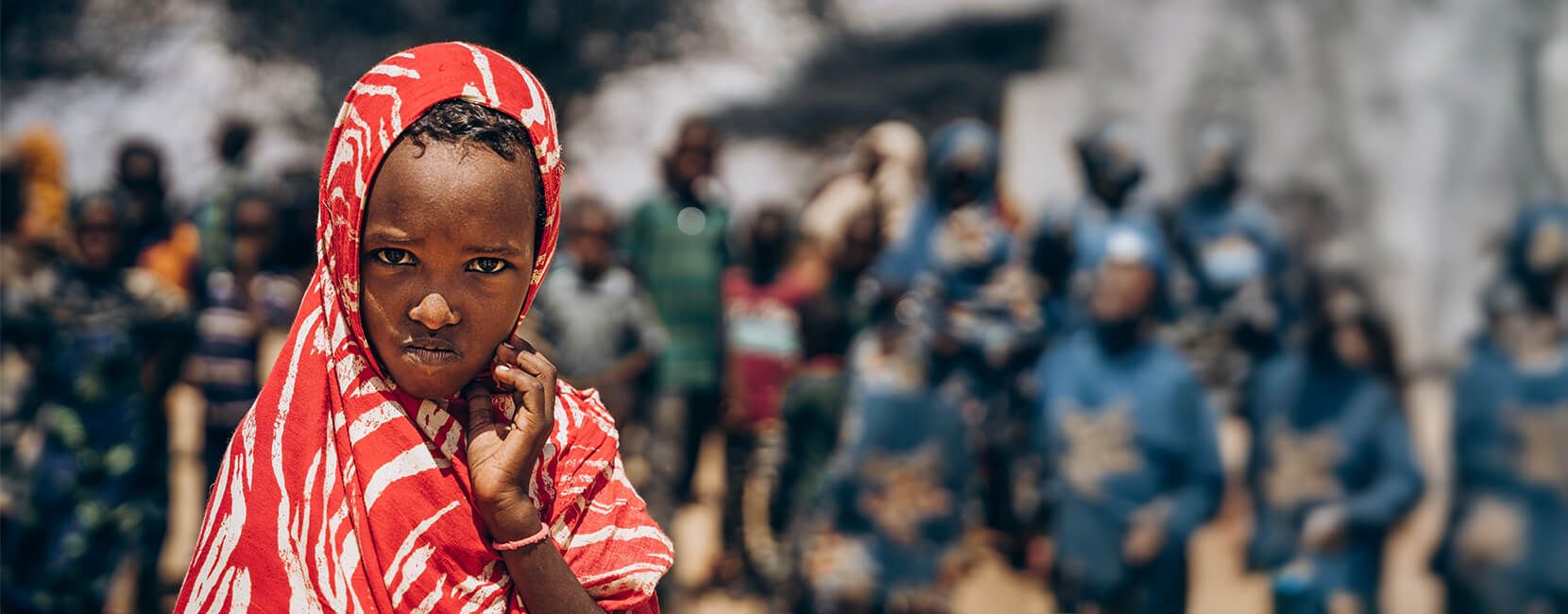 A young girl in a red and white patterned headscarf stands in focus, looking at the camera with a serious expression, with a blurred crowd in the background.