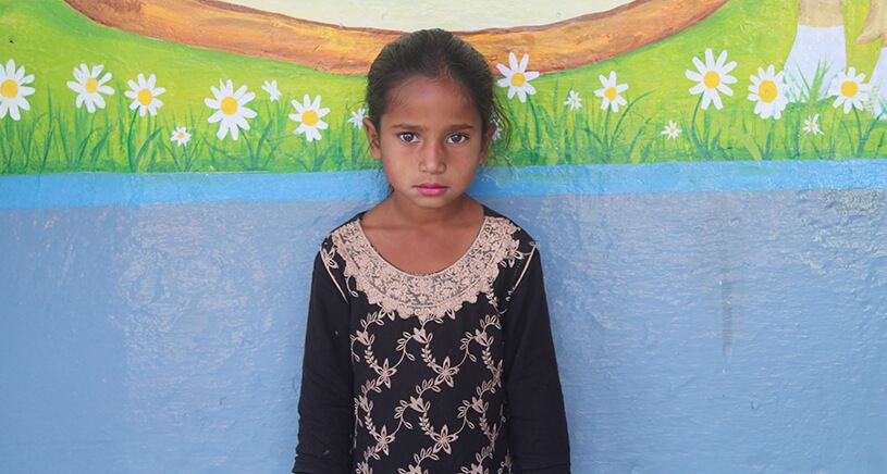 A young girl with a serious expression stands against a colorful wall mural featuring grass, flowers, and a painted tree. She is wearing a black dress with beige embroidery.