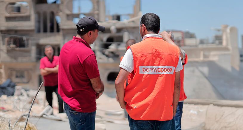 Aid workers in orange vests from Penny Appeal converse in front of a damaged building, with rubble scattered around.