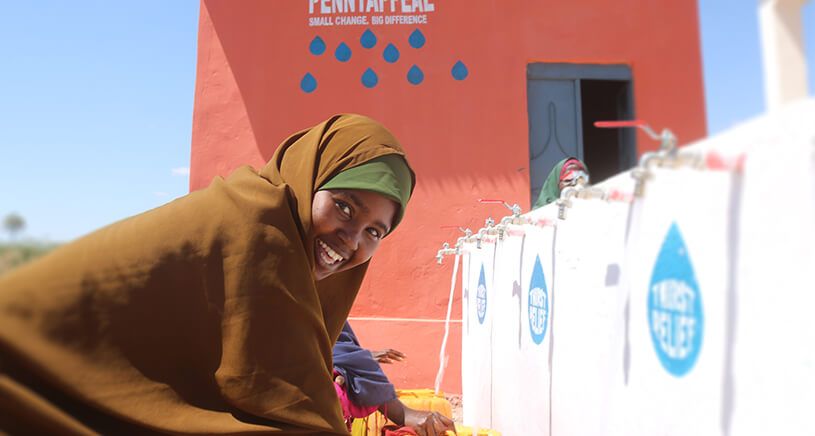 A woman smiles while collecting water from a community water point in a rural area. She is wearing a brown hijab and green headscarf, with a bright orange water tower in the background, featuring the Penny Appeal Thirst Relief logo.