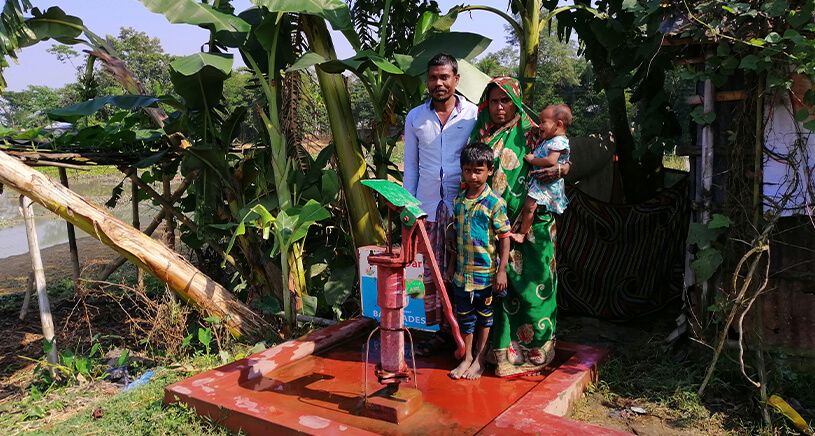 A family of five stands around a hand pump in a rural setting, surrounded by banana trees and greenery. The father, mother holding a baby, and two children stand on a red cement platform with the pump in the center.