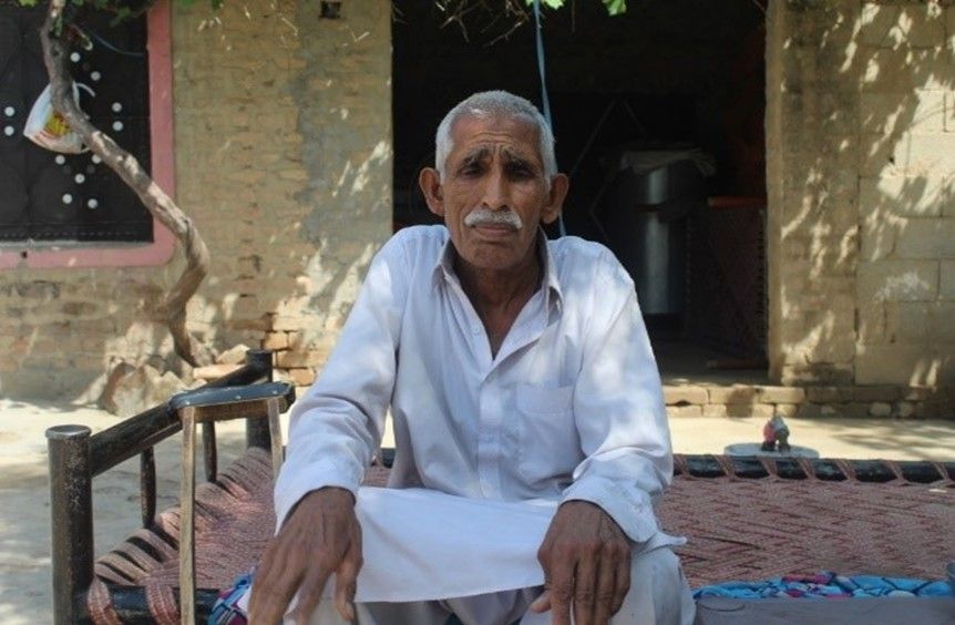 An elderly man sitting on a traditional woven bed (charpai) outdoors, wearing a white kurta. He has a mustache and is looking directly at the camera, with a neutral expression.