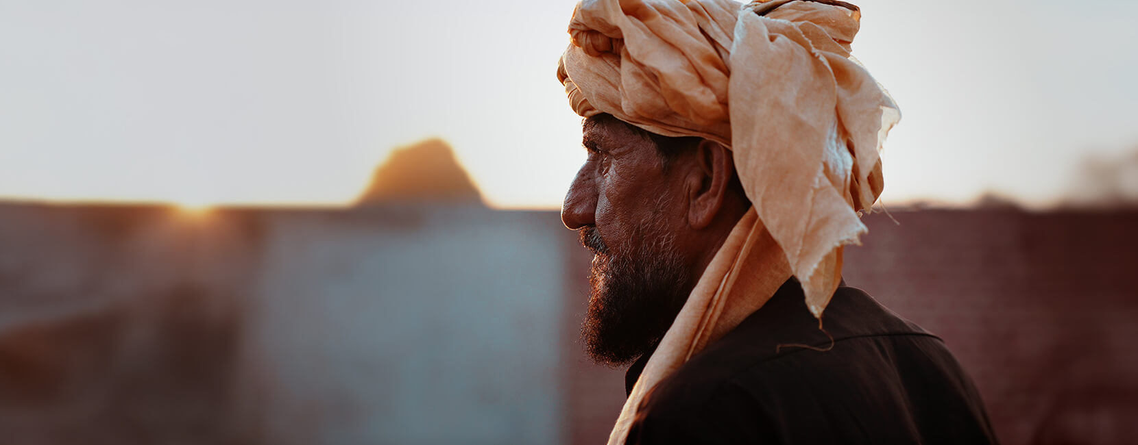 Side profile of an elderly man wearing a traditional turban, standing outdoors at sunset with a calm expression, highlighting dignity and resilience.